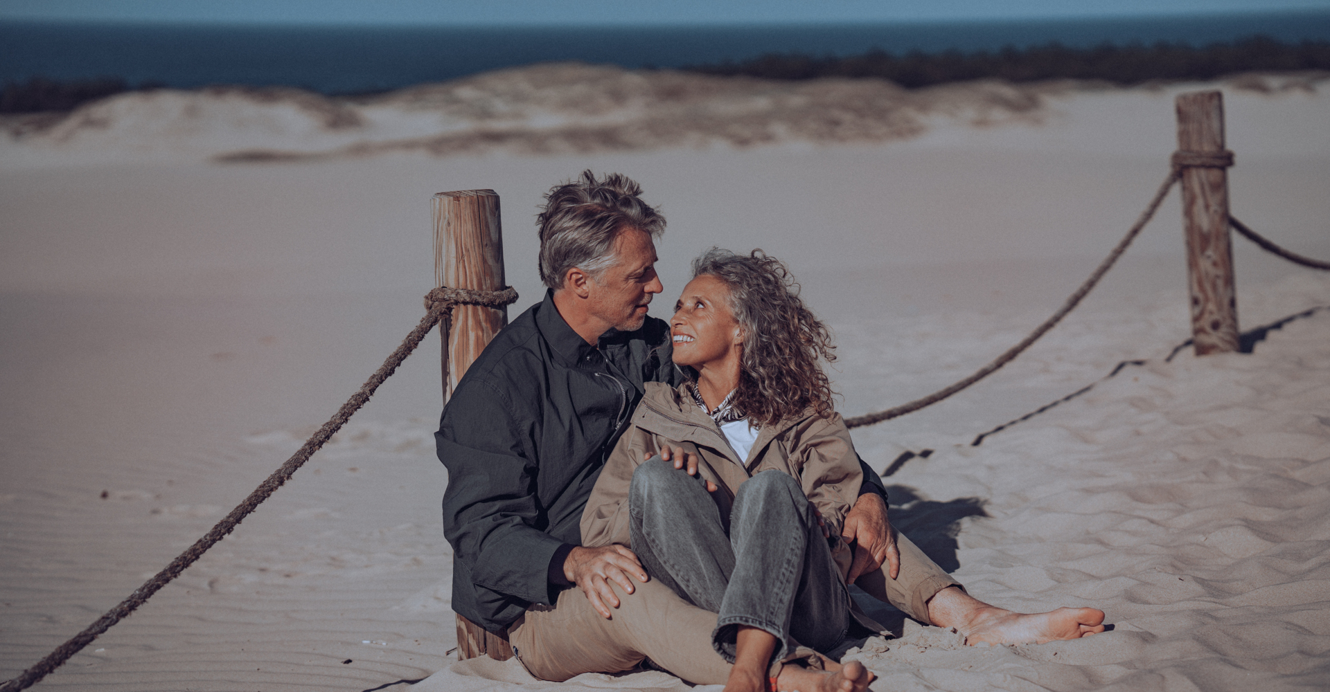 Mature couple seated on sandy dunes near rope posts, smiling and embracing with ocean horizon in background