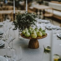 Elegant dining table set for an event with white linens, wine glasses, rattan chairs, wooden bowl of fresh pears and leafy centerpiece