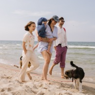 Family walking barefoot along a sunny sandy beach with small waves, mom carrying a toddler, smiling kids and a dog