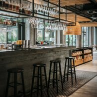 Modern hotel bar with concrete counter, hanging wine glasses, backlit bottles, wooden slatted ceiling and woven pendant lamps