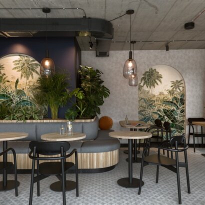 Cozy hotel lounge-café with curved banquette seating, round wooden tables, pendant lights, tropical mural arches and lush plants