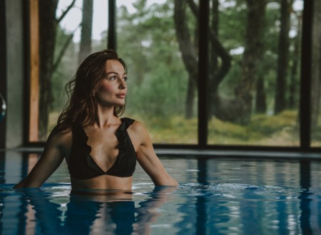 Woman relaxing in an indoor hotel pool by floor-to-ceiling windows overlooking a forest, spa-like calm ambiance