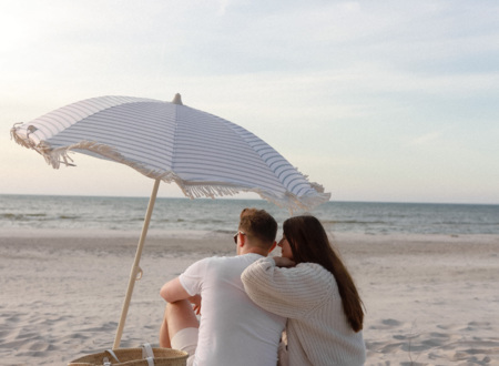 Couple cuddling on a blanket under a striped beach umbrella, straw tote beside them, calm sea at sunset