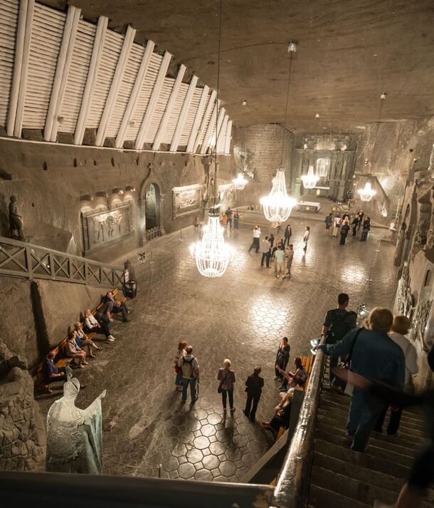 Podziemna komnata solna z kryształowymi żyrandolami, rzeźbami i płaskorzeźbami w soli, ławkami i zwiedzającymi w rozległej sali.