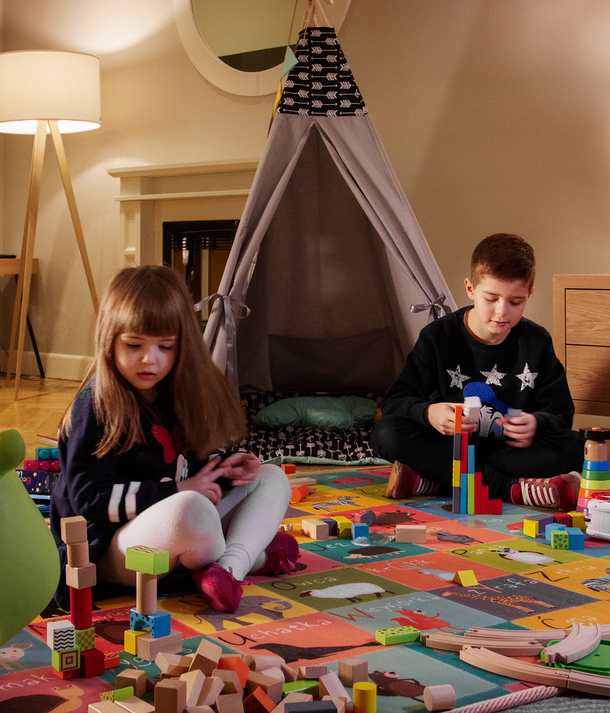 Dzieci bawią się w przytulnej rodzinnej sali zabaw z tipi, kolorową matą, drewnianymi klockami i ciepłym oświetleniem.