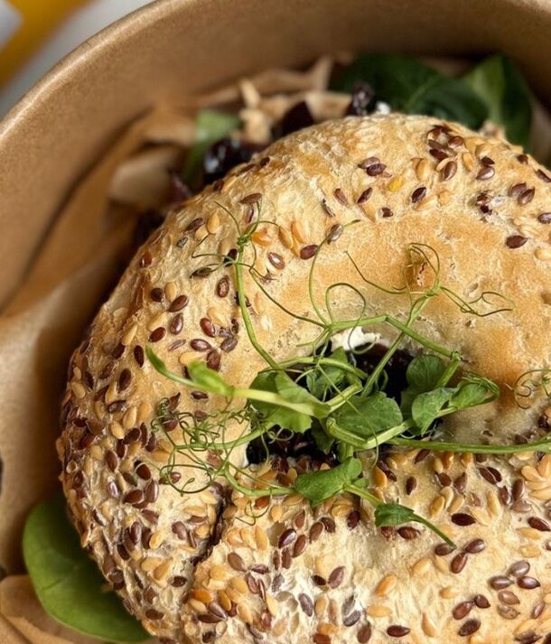 Bagel z ziarnami, z sałatą i kiełkami w brązowej misce na wynos, obok butelka soku pomarańczowego