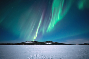 Grüne Polarlichter über einem zugefrorenen See und bewaldeten Hügeln — perfekter Ort zur Nordlichtbeobachtung