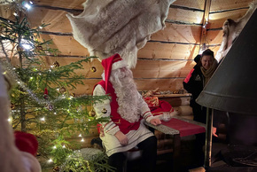 Santa in red suit seated in a cozy wooden cabin beside a lit Christmas tree, fur wall hide, wood stove and smiling guest