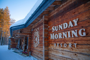 Rustic log-resort entrance with snow-covered roof, warm wooden façade reading 'SUNDAY MORNING RESORT' and decorated porch by pine trees