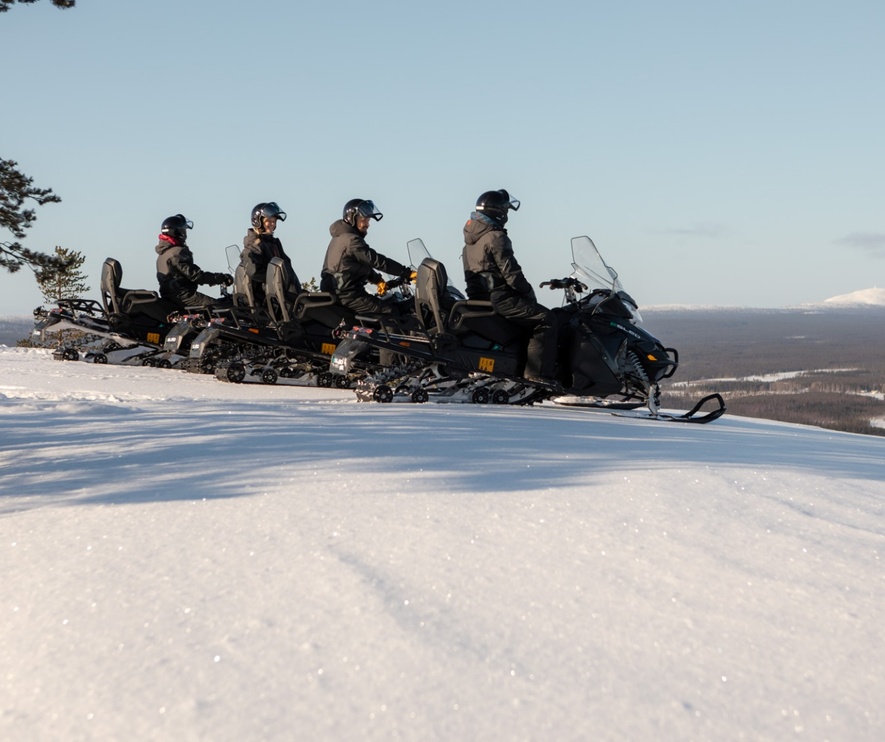 Guided snowmobile tour on a snowy ridge with four riders in winter gear overlooking a vast frozen forest